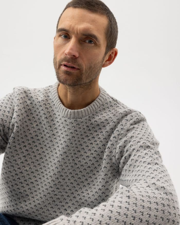 A man with short brown hair and a trimmed beard wears a light gray sweater with a small, repeating dark pattern, looking slightly upward against a plain, light background.