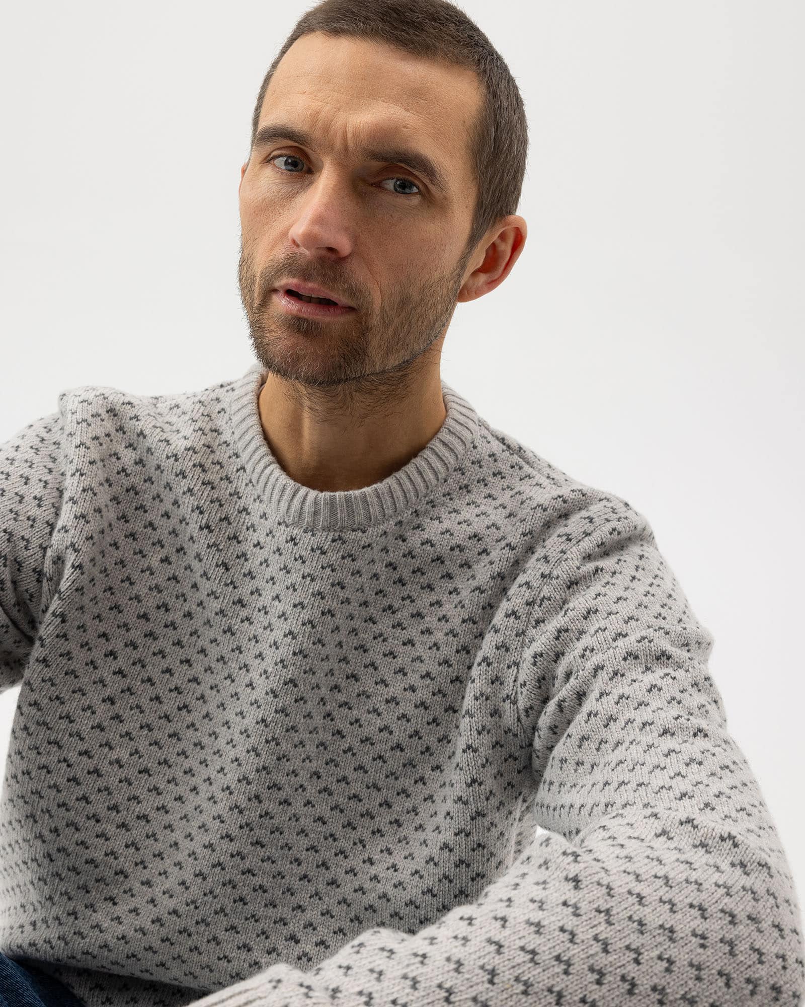 A man with short brown hair and a trimmed beard wears a light gray sweater with a small, repeating dark pattern, looking slightly upward against a plain, light background.