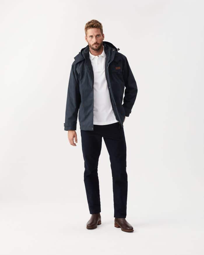 A man with short hair and a beard stands against a white background in an R M Williams Rockley Jacket, white polo shirt, dark pants, and brown boots, one hand in his pocket and wearing a neutral expression.
