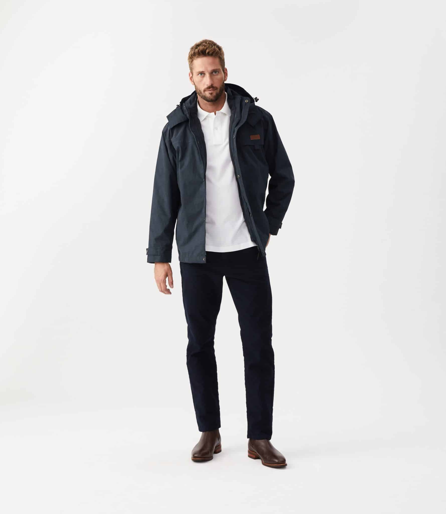 A man with short hair and a beard stands against a white background in an R M Williams Rockley Jacket, white polo shirt, dark pants, and brown boots, one hand in his pocket and wearing a neutral expression.