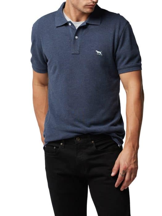 A man wearing a short-sleeve, dark blue polo shirt with a small animal logo on the chest and black jeans, standing with one hand in his pocket.