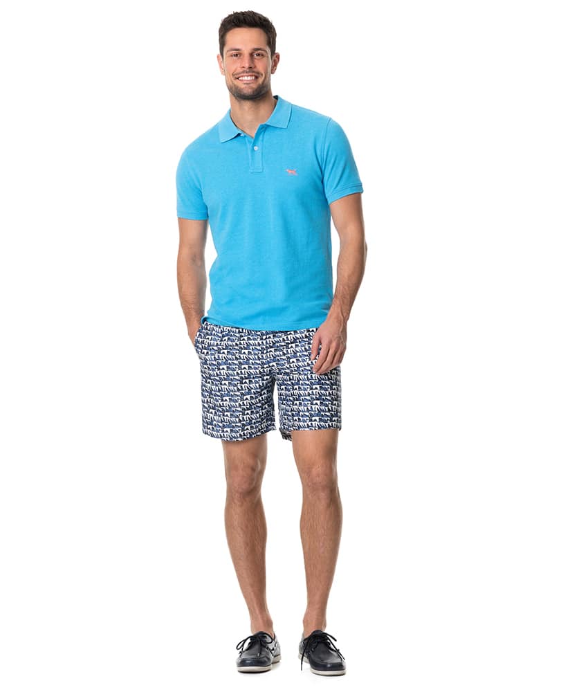 A smiling man stands with hands in pockets, wearing a bright blue polo shirt, patterned blue and white shorts, and dark boat shoes. He faces forward against a plain white background.