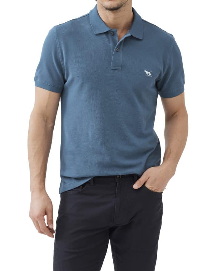 A man wearing a teal short-sleeve polo shirt with a small embroidered logo on the chest and dark pants, standing with one hand in his pocket against a white background.
