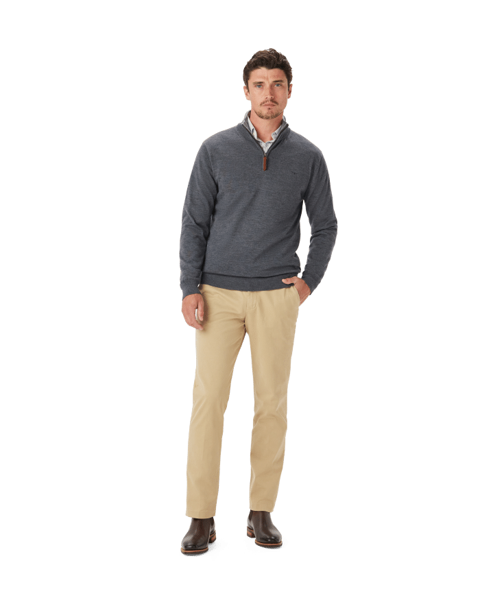 A man stands against a white background, wearing a dark blue quarter-zip sweater over a collared shirt, beige pants, and brown boots. He has one hand in his pocket and a neutral expression.