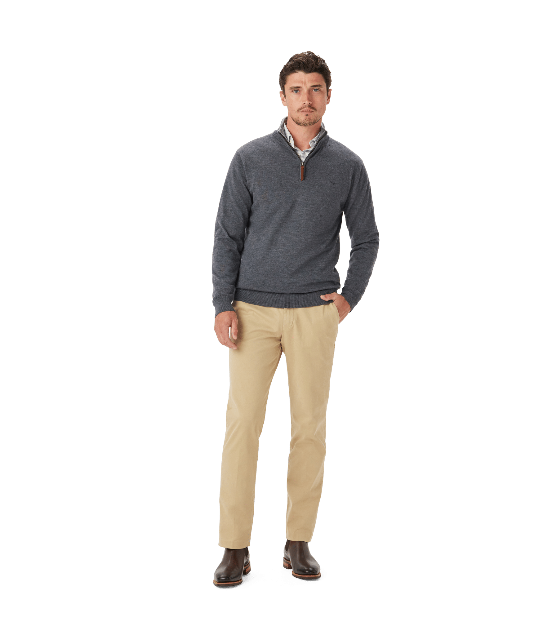 A man stands against a white background, wearing a dark blue quarter-zip sweater over a collared shirt, beige pants, and brown boots. He has one hand in his pocket and a neutral expression.