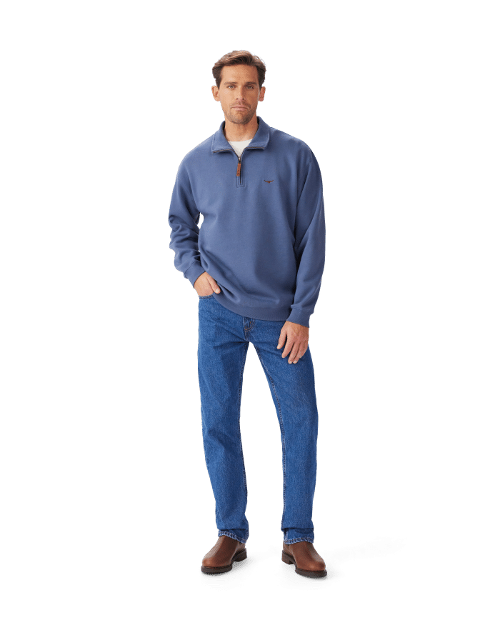 A man stands against a white background wearing a blue quarter-zip pullover, blue jeans, and brown shoes, with one hand in his pocket and a neutral expression on his face.