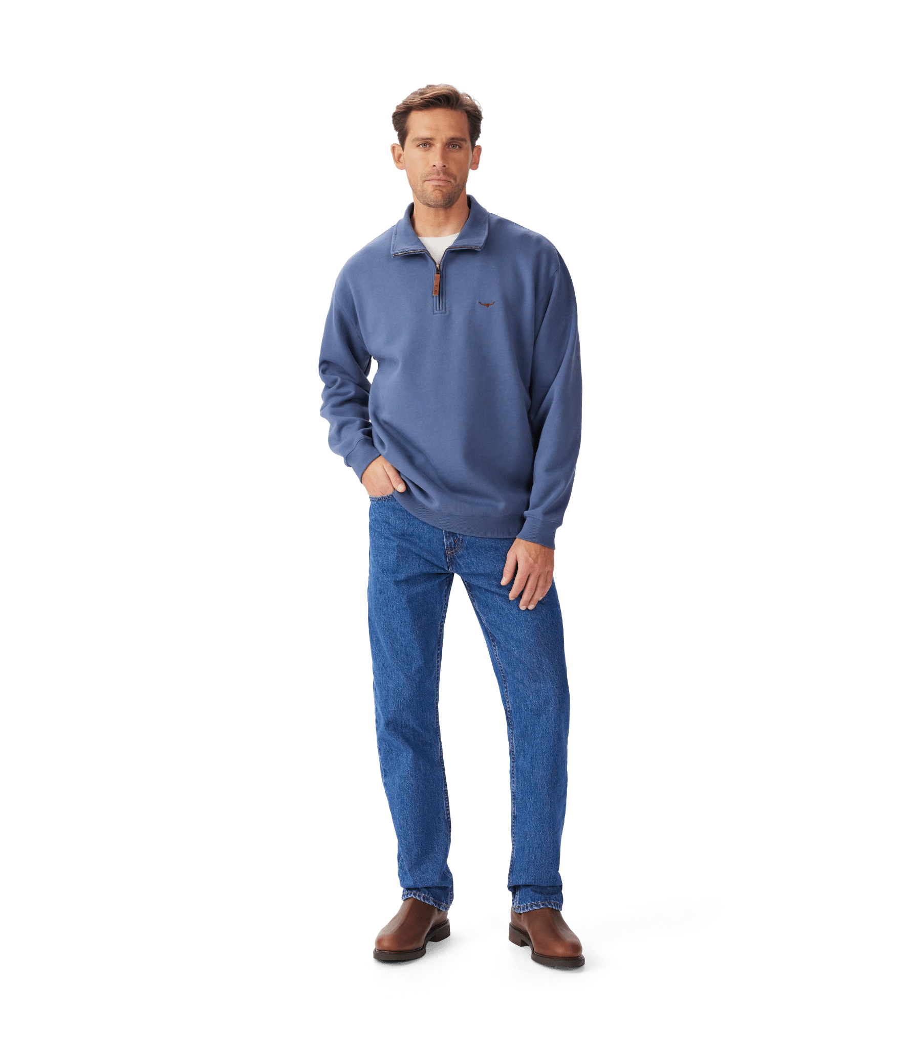 A man stands against a white background wearing a blue quarter-zip pullover, blue jeans, and brown shoes, with one hand in his pocket and a neutral expression on his face.