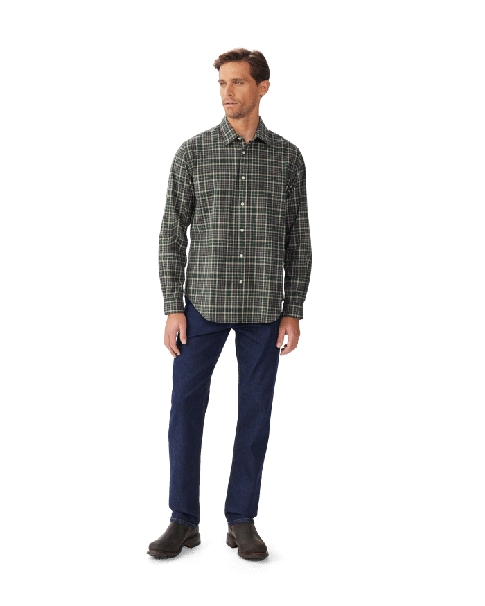 A man stands against a plain white background wearing a green plaid button-up shirt, dark blue jeans, and brown shoes. He is looking slightly to his left with a neutral expression.