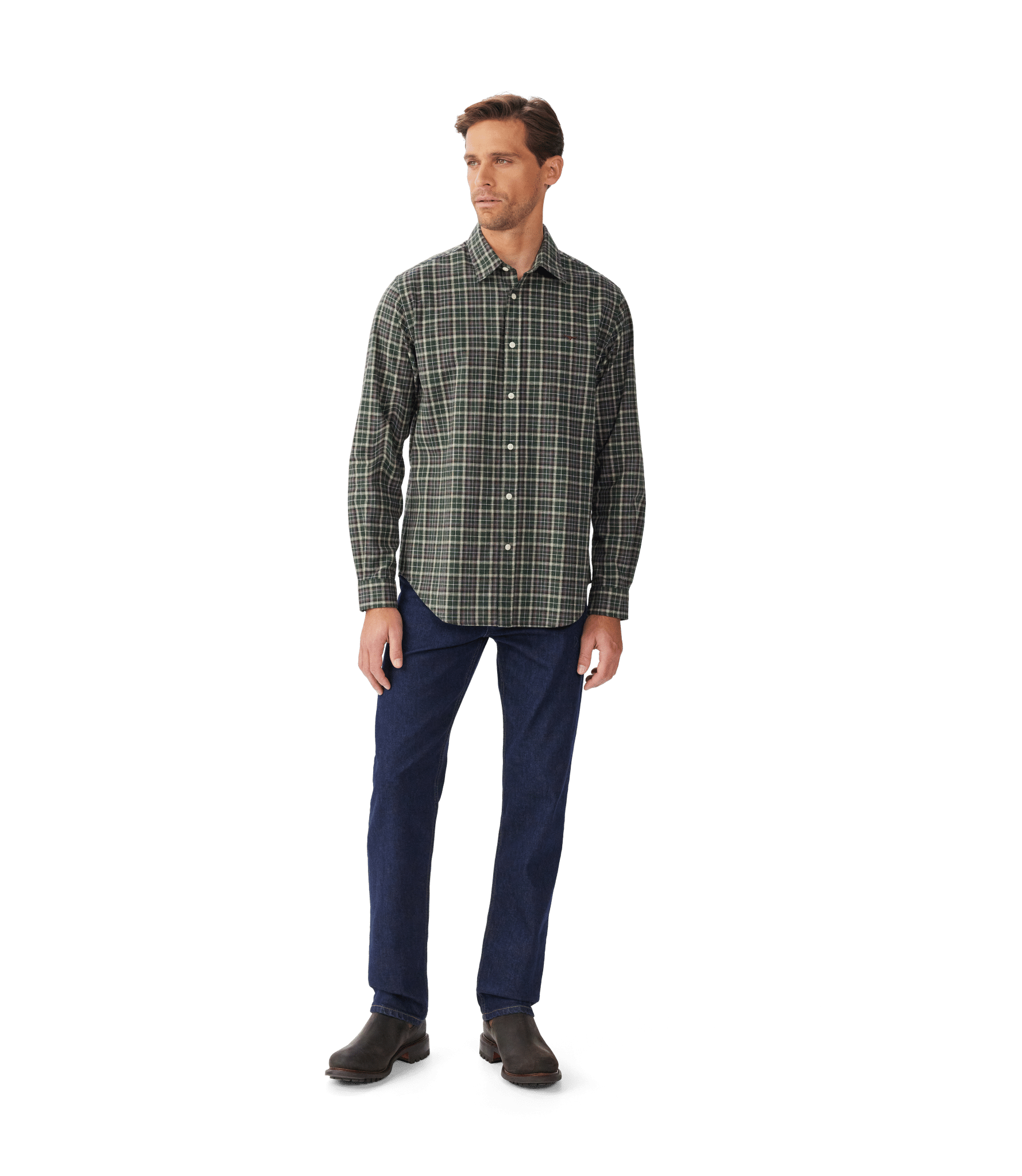 A man stands against a plain white background wearing a green plaid button-up shirt, dark blue jeans, and brown shoes. He is looking slightly to his left with a neutral expression.