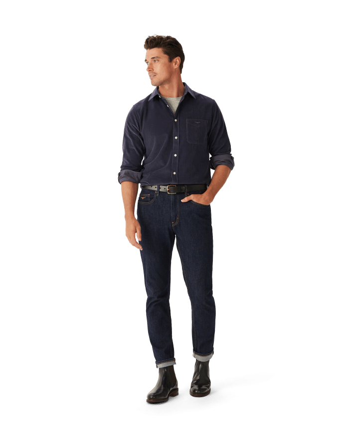 A man stands with one hand in his pocket, wearing a navy button-up shirt over a gray t-shirt, dark blue cuffed jeans, a black belt, and black boots against a plain white background.