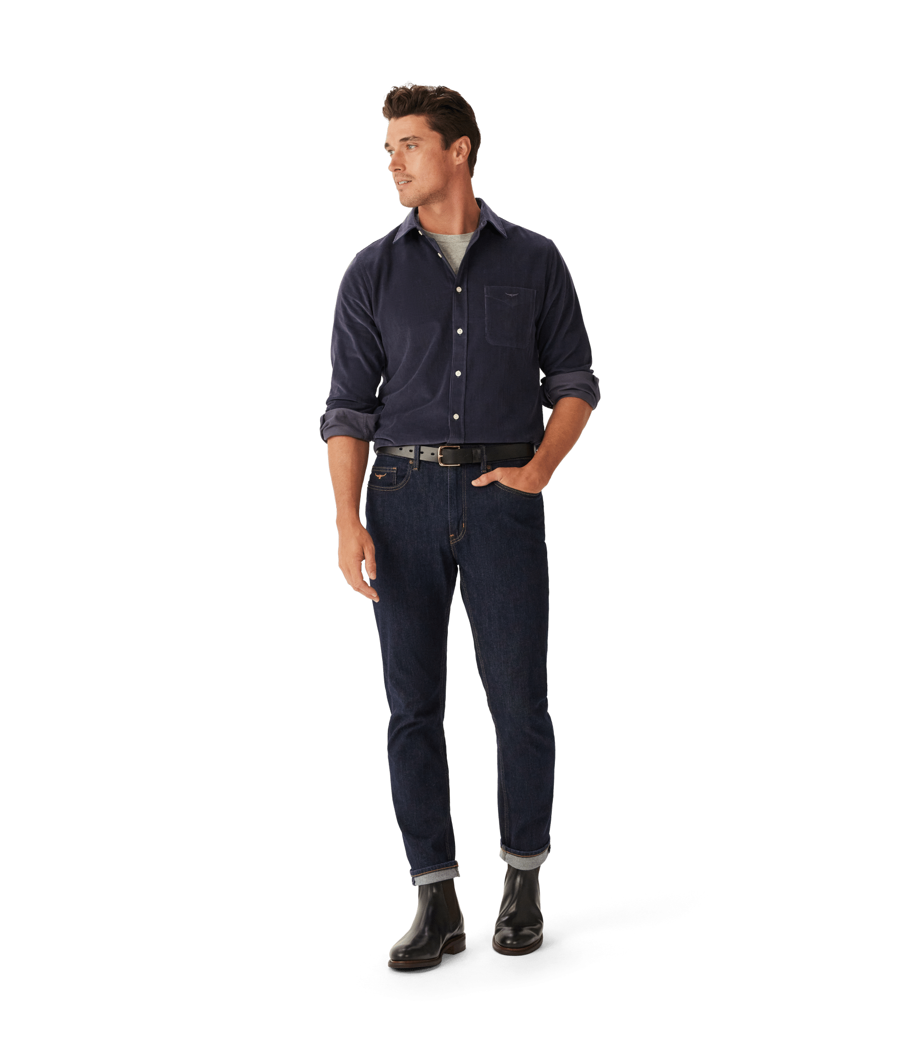 A man stands with one hand in his pocket, wearing a navy button-up shirt over a gray t-shirt, dark blue cuffed jeans, a black belt, and black boots against a plain white background.