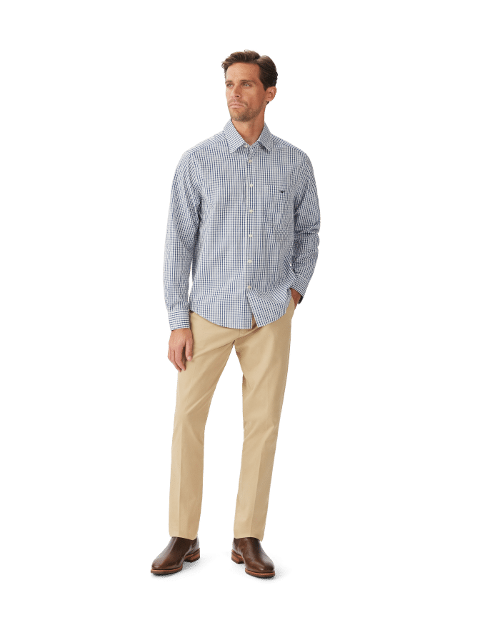 A man stands against a white background wearing a blue and white checkered button-down shirt, beige pants, and brown dress shoes. He has short hair and is looking slightly to his right.
