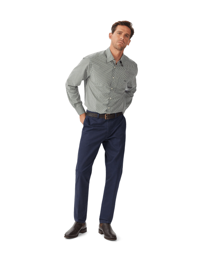 A man stands against a white background wearing a light gray checkered long-sleeve shirt, navy blue pants, a black belt, and dark brown shoes, with his hands in his pockets and a neutral expression.