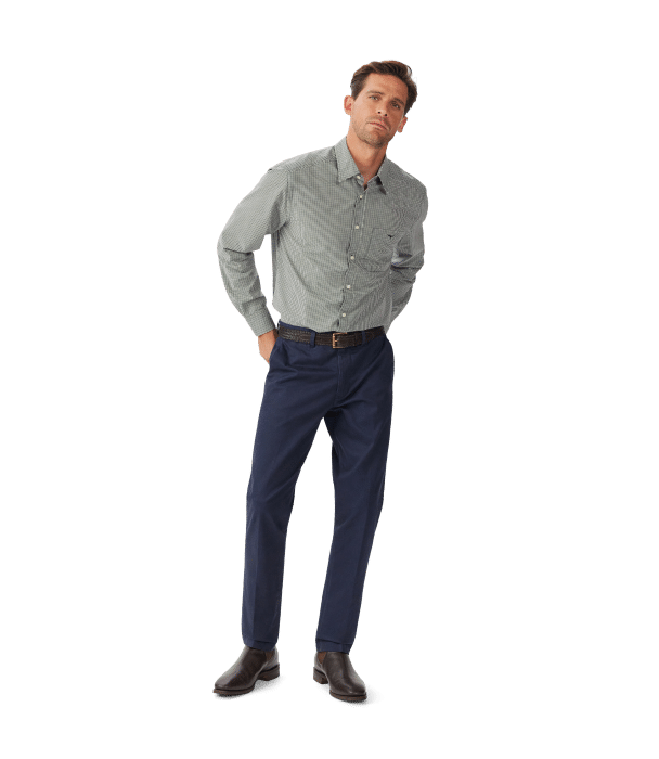 A man stands against a plain white background wearing a gray checkered button-up shirt, navy blue trousers, a brown belt, and brown dress shoes. His hands are in his pockets and he is looking slightly to the side.