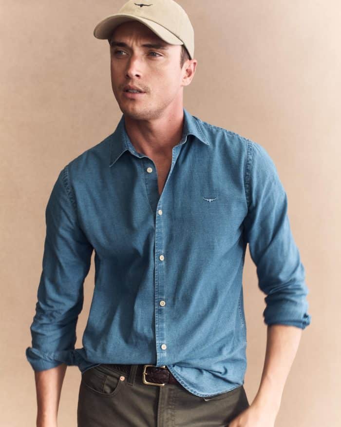 A man models the R M Williams Arno Regular Denim Shirt with rolled sleeves, paired with dark pants and a beige cap, standing against a beige background and gazing to the side with a neutral expression.