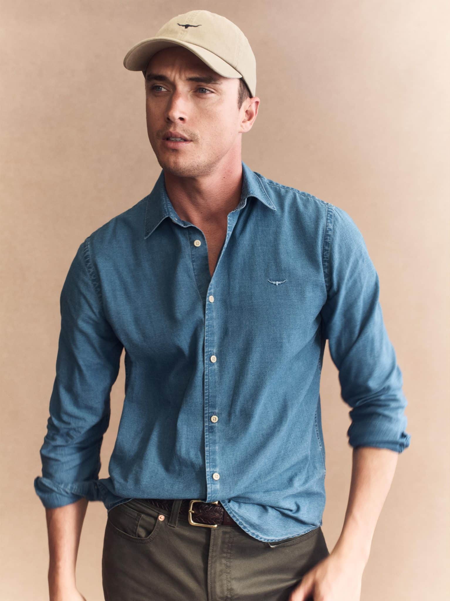 A man models the R M Williams Arno Regular Denim Shirt with rolled sleeves, paired with dark pants and a beige cap, standing against a beige background and gazing to the side with a neutral expression.