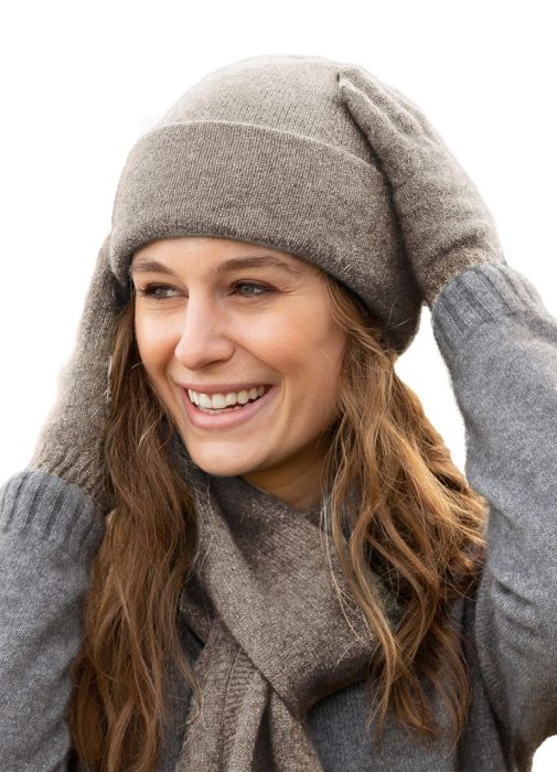A smiling woman wearing a gray knit hat, scarf, and gloves looks to the side. Her long brown hair is loose, and she appears happy and cozy against a plain white background.