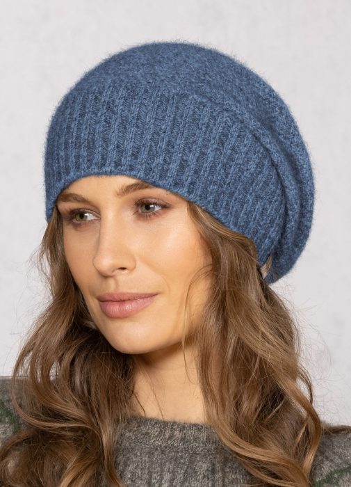 A woman with wavy brown hair wears a blue knitted slouchy beanie and a gray sweater, looking slightly to the side against a light, neutral background.