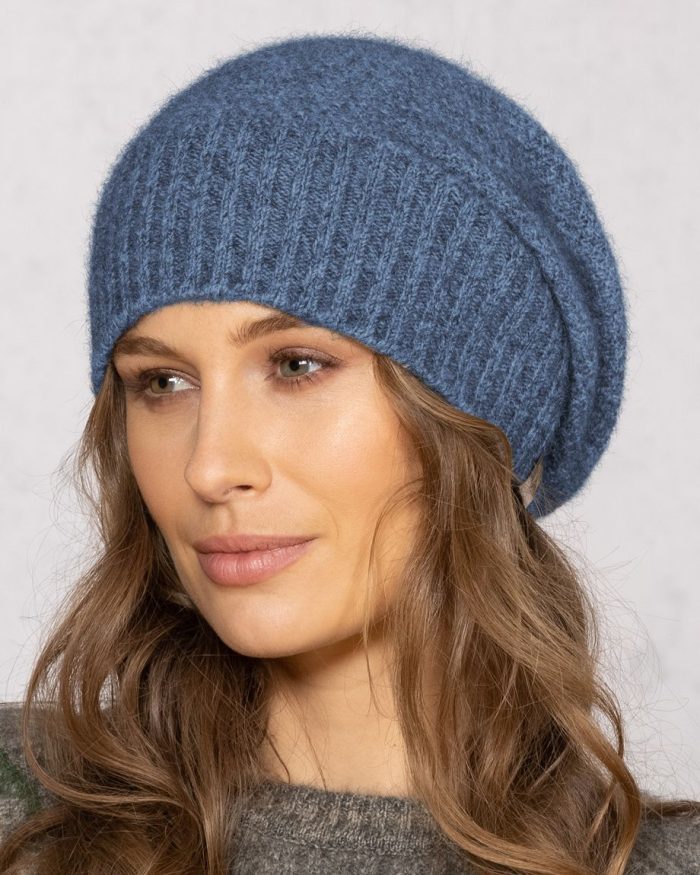 A woman with wavy brown hair wears a blue knitted slouchy beanie and a gray sweater, looking slightly to the side against a light, neutral background.