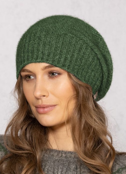 A woman with wavy brown hair wears a ribbed, dark green knit beanie and a grey sweater, looking slightly to the side with a neutral expression against a light background.