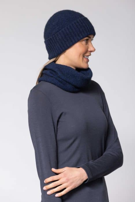 A woman in a navy knit hat and matching scarf looks to the side, smiling. She is wearing a long-sleeve dark blue top against a plain light background.
