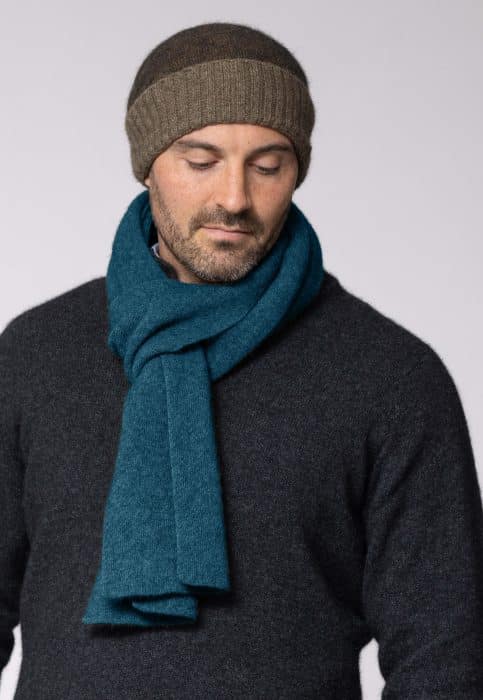 A man wearing a brown knit beanie, a dark gray sweater, and a teal scarf looks down with a neutral expression against a plain light background.