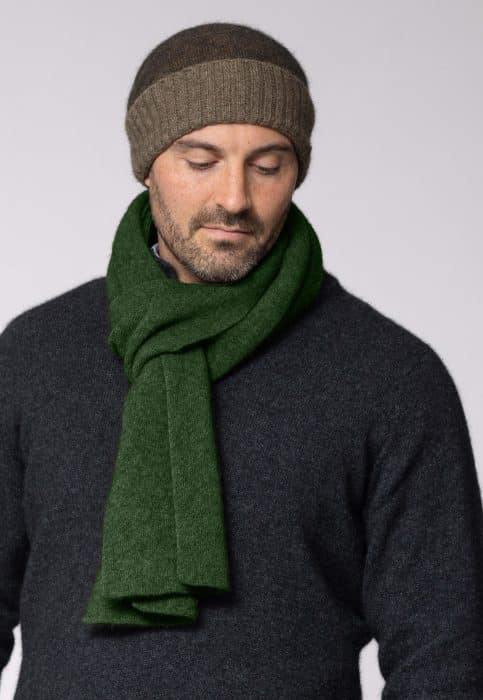 A man wearing a brown knit beanie, a dark charcoal sweater, and a thick green scarf looks down with a neutral expression against a plain light background.