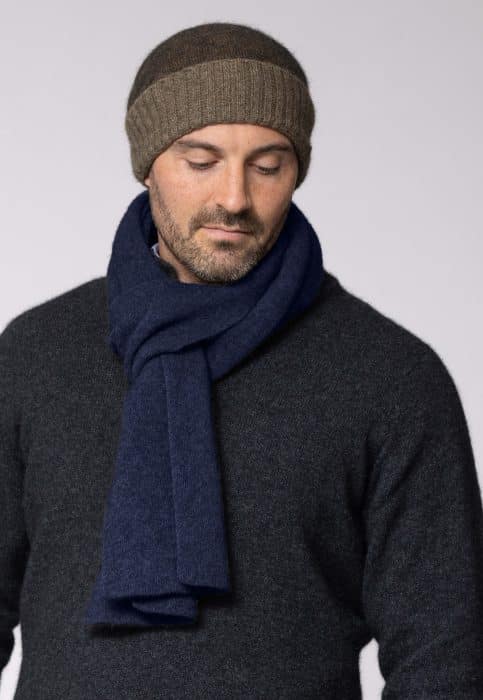 A man wearing a brown knit beanie, a dark grey sweater, and a navy blue scarf looks down with a neutral expression against a plain light background.