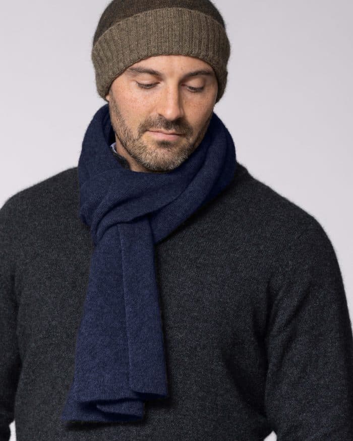 A man wearing a brown knit beanie, a dark grey sweater, and a navy blue scarf looks down with a neutral expression against a plain light background.