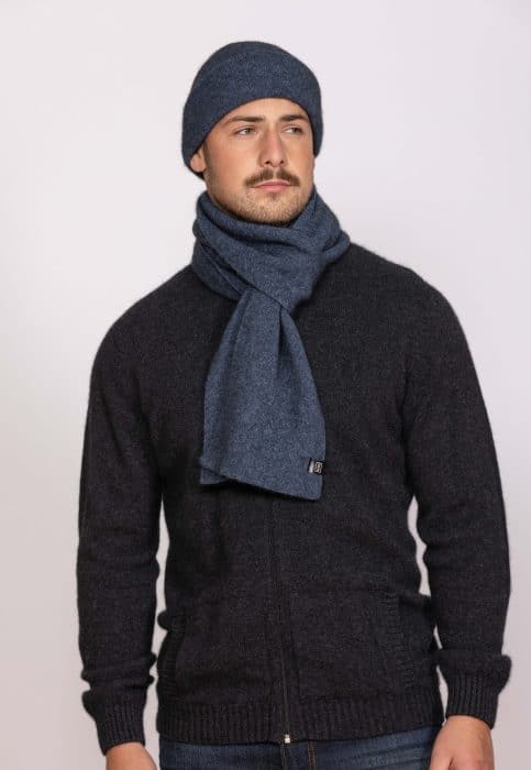 A man wearing a dark zip-up sweater, a blue knit scarf, and a matching blue knit beanie stands against a plain light background, looking slightly to the side with a neutral expression.