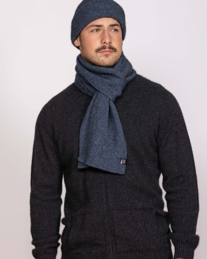 A man wearing a dark zip-up sweater, a blue knit scarf, and a matching blue knit beanie stands against a plain light background, looking slightly to the side with a neutral expression.