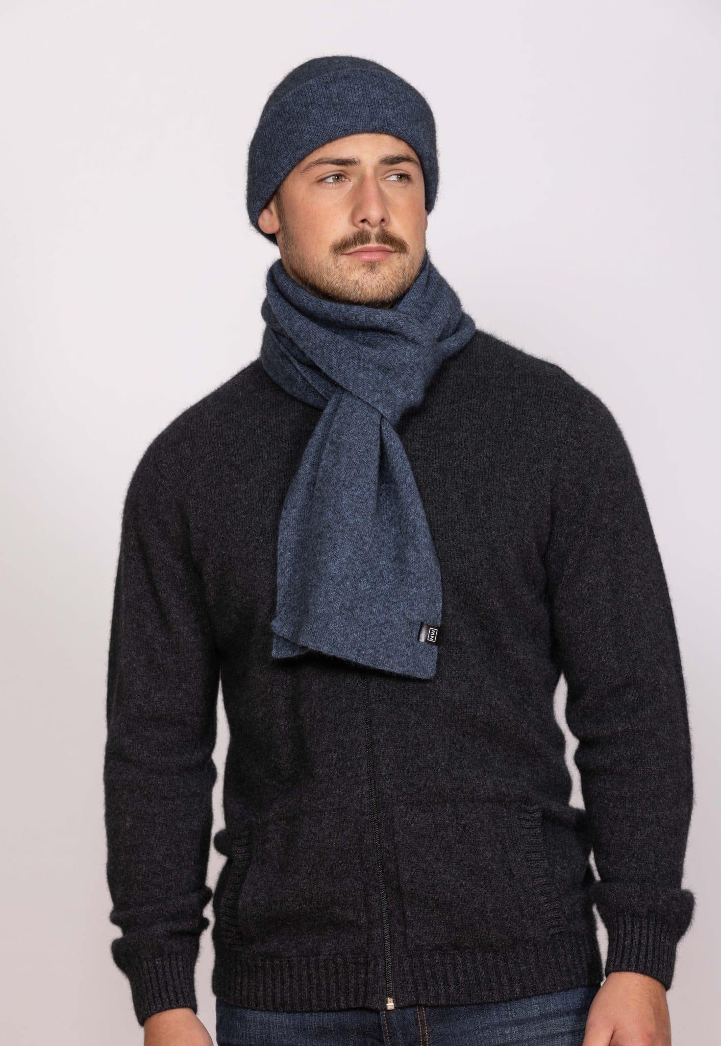 A man wearing a dark zip-up sweater, a blue knit scarf, and a matching blue knit beanie stands against a plain light background, looking slightly to the side with a neutral expression.