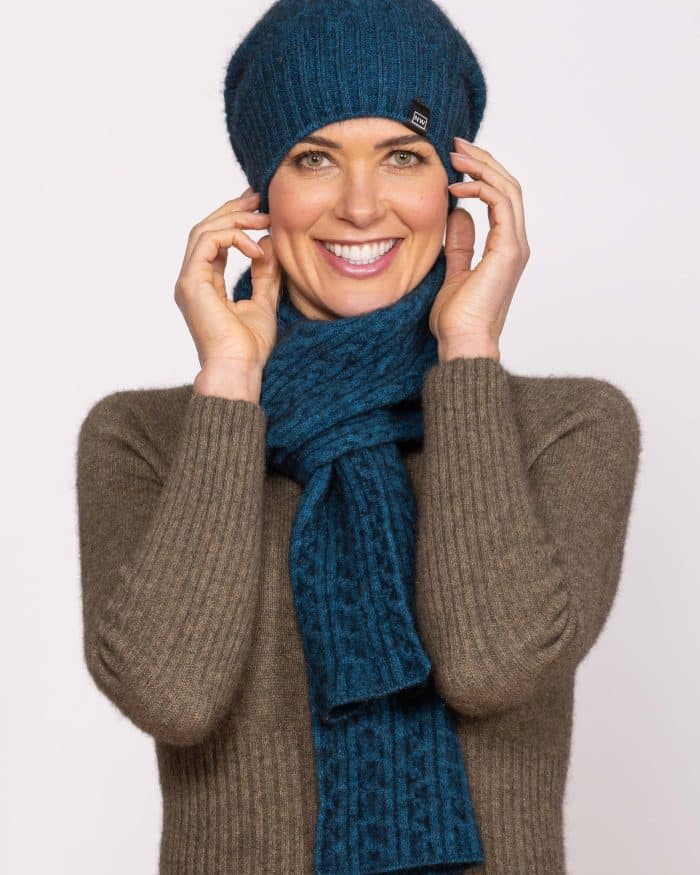 A smiling woman wears a matching blue knit hat and scarf, along with a brown ribbed sweater, holding the sides of her hat with both hands against a plain light background.