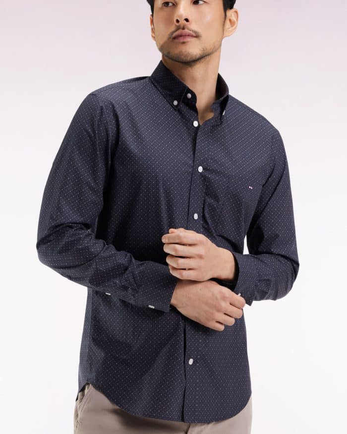 A man wearing a dark navy button-up shirt with small white polka dots and light beige pants stands against a light gradient background, looking slightly to the side.