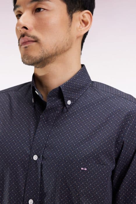 A man with short dark hair and a trimmed beard wears a dark patterned button-up shirt with a small embroidered logo on the chest pocket, shown from the shoulders up against a light gradient background.