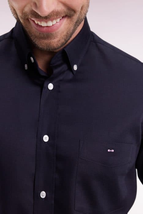 A man wearing a dark navy button-up shirt with white buttons, smiling. The shirt has a front pocket on the left side with a small embroidered logo. His face is partially visible, focusing on the mouth and chin.
