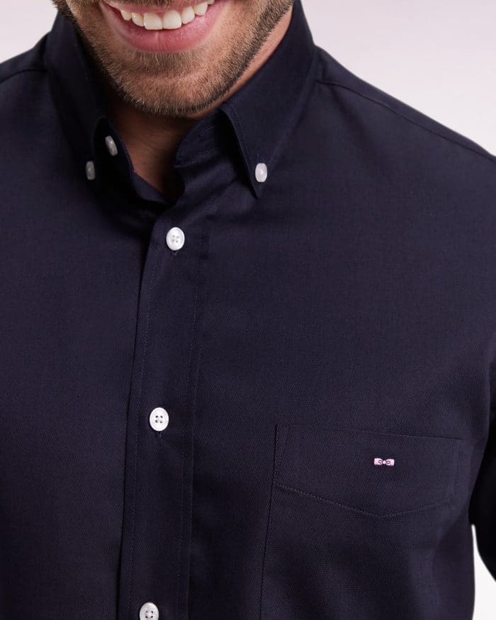 A man wearing a dark navy button-up shirt with white buttons, smiling. The shirt has a front pocket on the left side with a small embroidered logo. His face is partially visible, focusing on the mouth and chin.