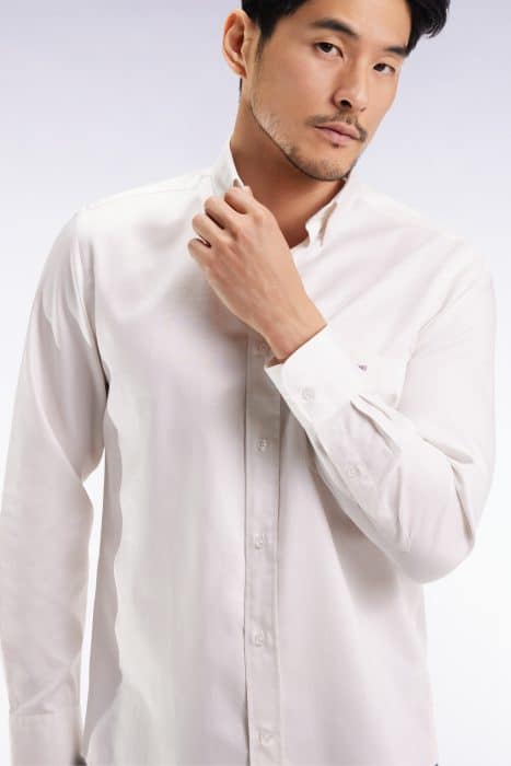 A man wearing a white long-sleeve button-up shirt adjusts his collar while looking slightly to the side against a plain, light background.