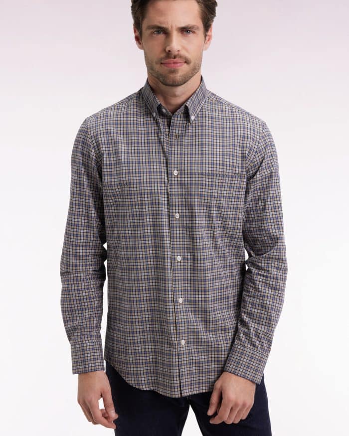 A man with light brown hair and a short beard is wearing a long-sleeve, button-up plaid shirt in shades of blue, brown, and white, standing against a light, gradient background.