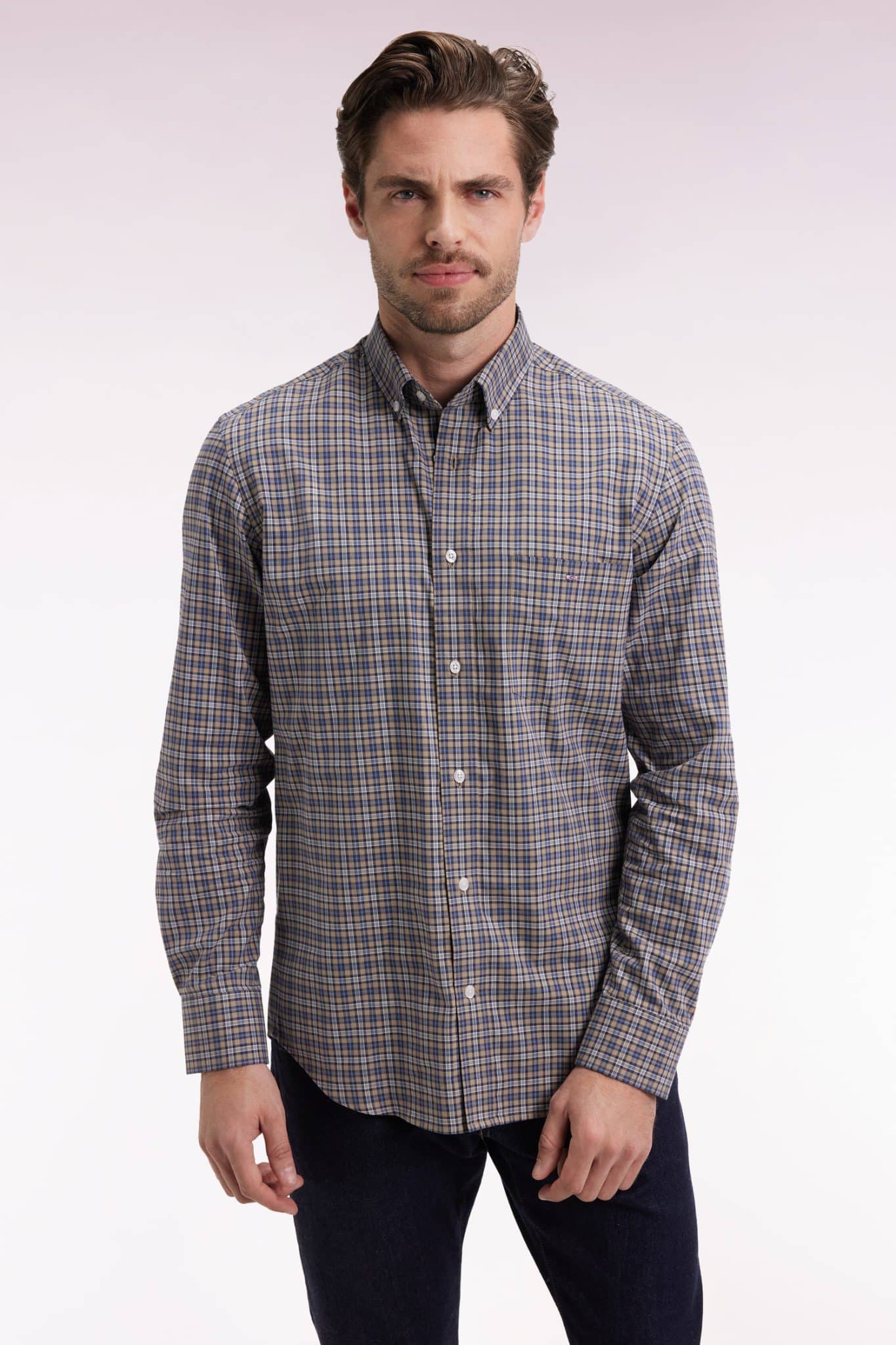 A man with light brown hair and a short beard is wearing a long-sleeve, button-up plaid shirt in shades of blue, brown, and white, standing against a light, gradient background.
