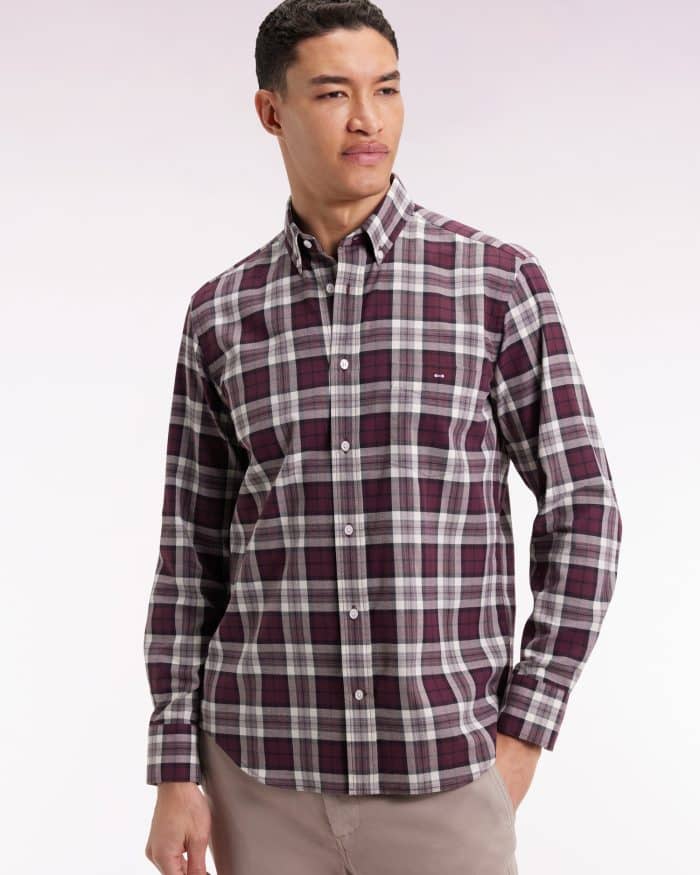 A man wearing a long-sleeve, button-up plaid shirt in shades of burgundy, white, and gray, paired with light beige pants, standing against a plain light background.
