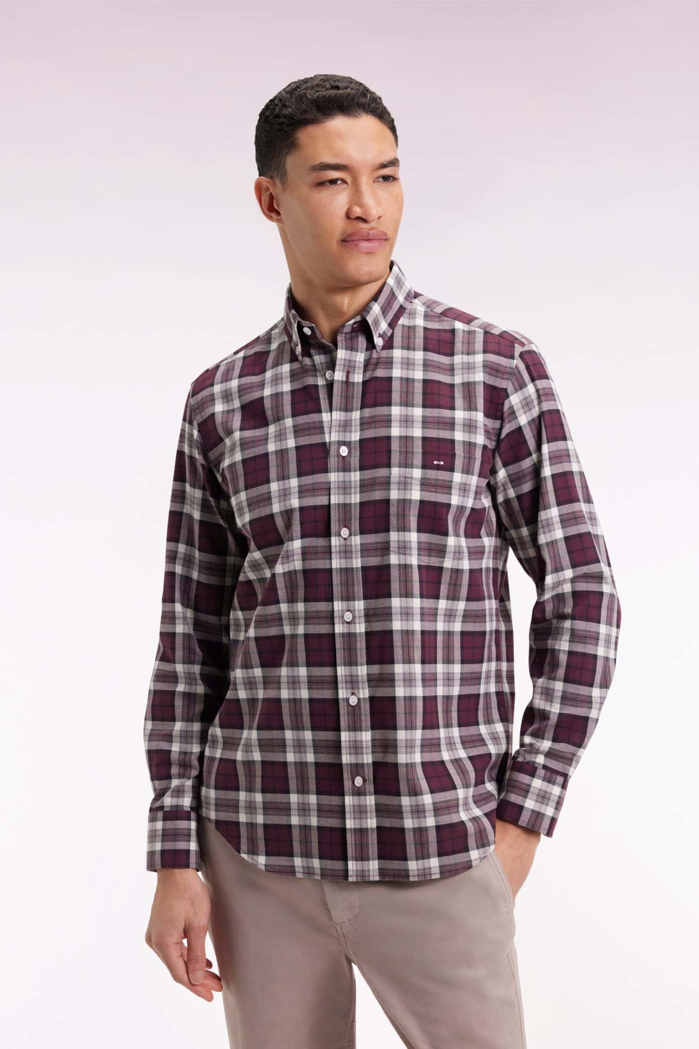 A man wearing a long-sleeve, button-up plaid shirt in shades of burgundy, white, and gray, paired with light beige pants, standing against a plain light background.