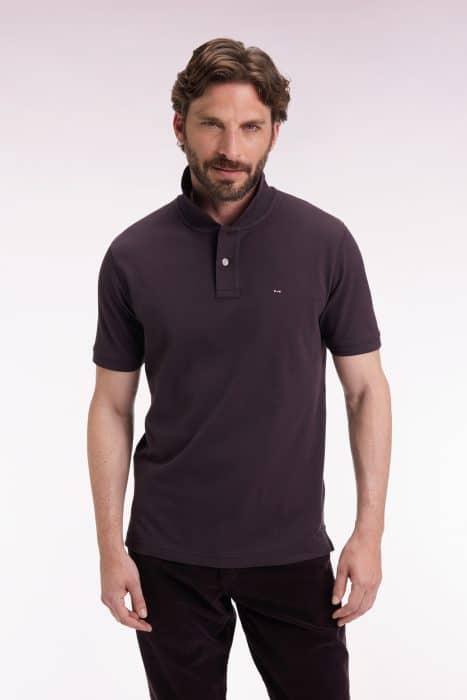 A man with brown hair and a beard is wearing a dark polo shirt and dark pants, standing against a plain, light background and looking at the camera with a slight smile.