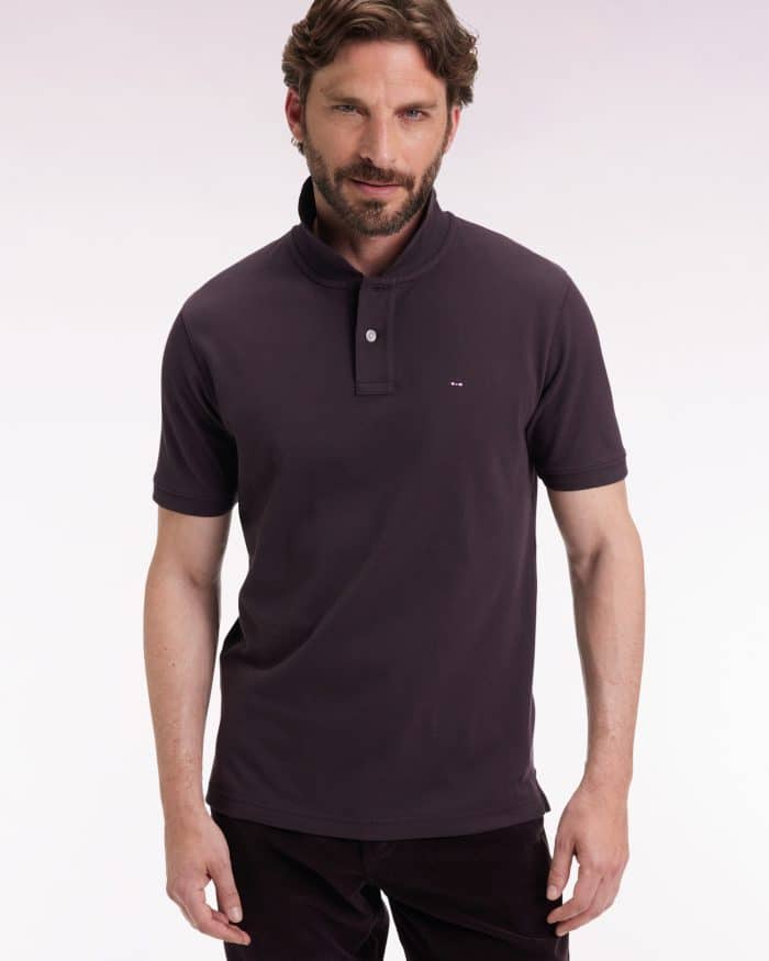 A man with brown hair and a beard is wearing a dark polo shirt and dark pants, standing against a plain, light background and looking at the camera with a slight smile.