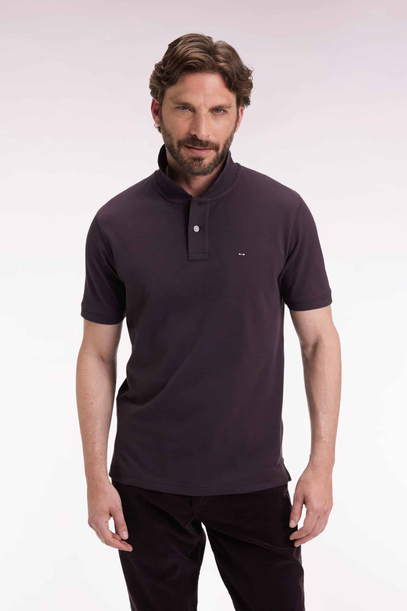 A man with brown hair and a beard is wearing a dark polo shirt and dark pants, standing against a plain, light background and looking at the camera with a slight smile.
