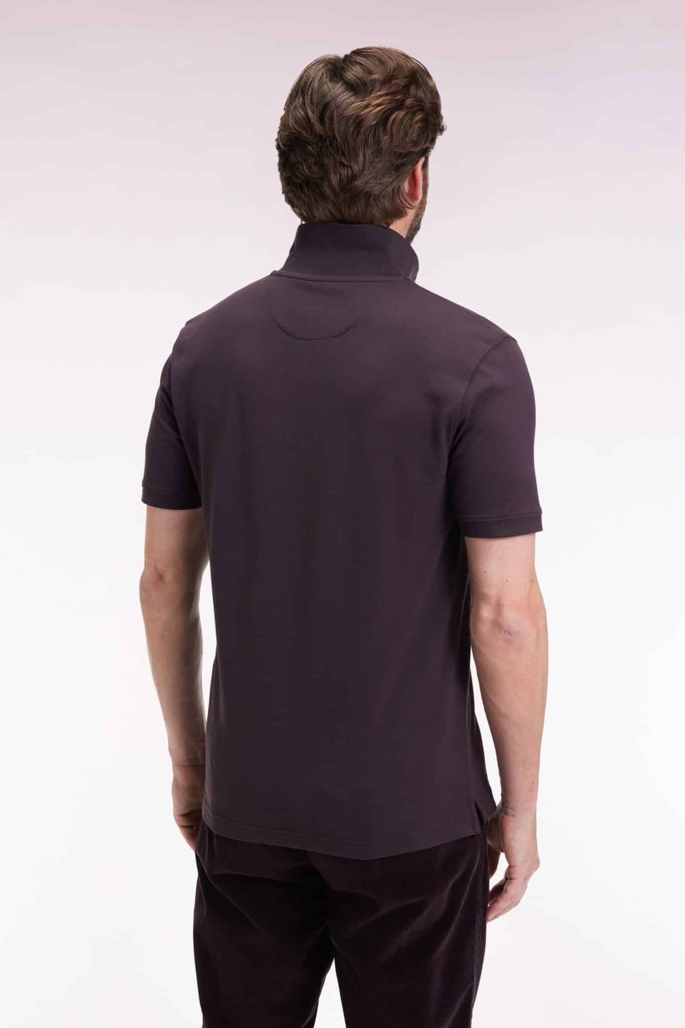 A man with brown hair is shown from the back wearing a short-sleeve, dark-colored polo shirt and dark pants against a light, plain background.