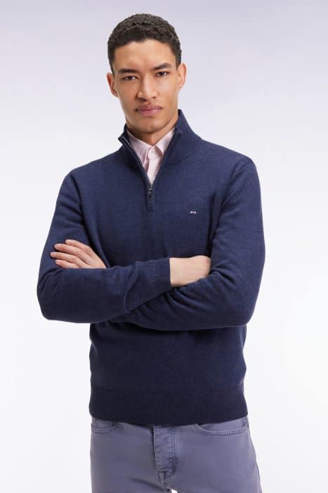 A man with short curly hair stands with arms crossed, wearing a navy quarter-zip sweater over a light pink collared shirt and gray pants, against a plain light background.