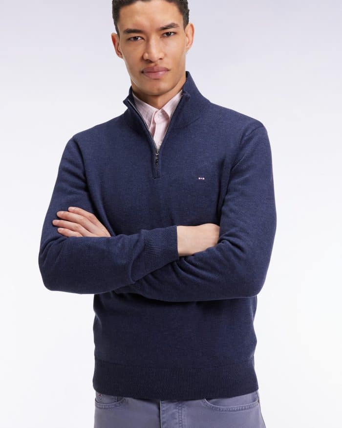 A man with short curly hair stands with arms crossed, wearing a navy quarter-zip sweater over a light pink collared shirt and gray pants, against a plain light background.