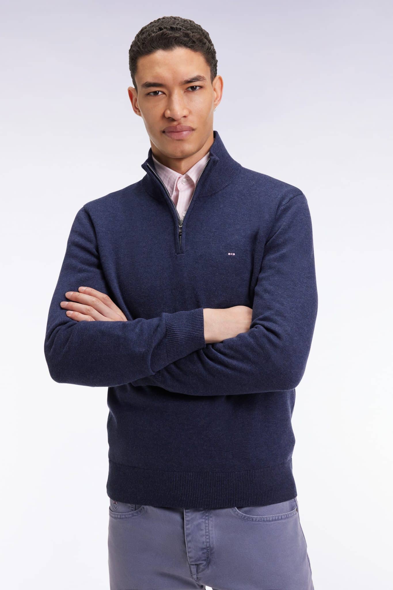 A man with short curly hair stands with arms crossed, wearing a navy quarter-zip sweater over a light pink collared shirt and gray pants, against a plain light background.