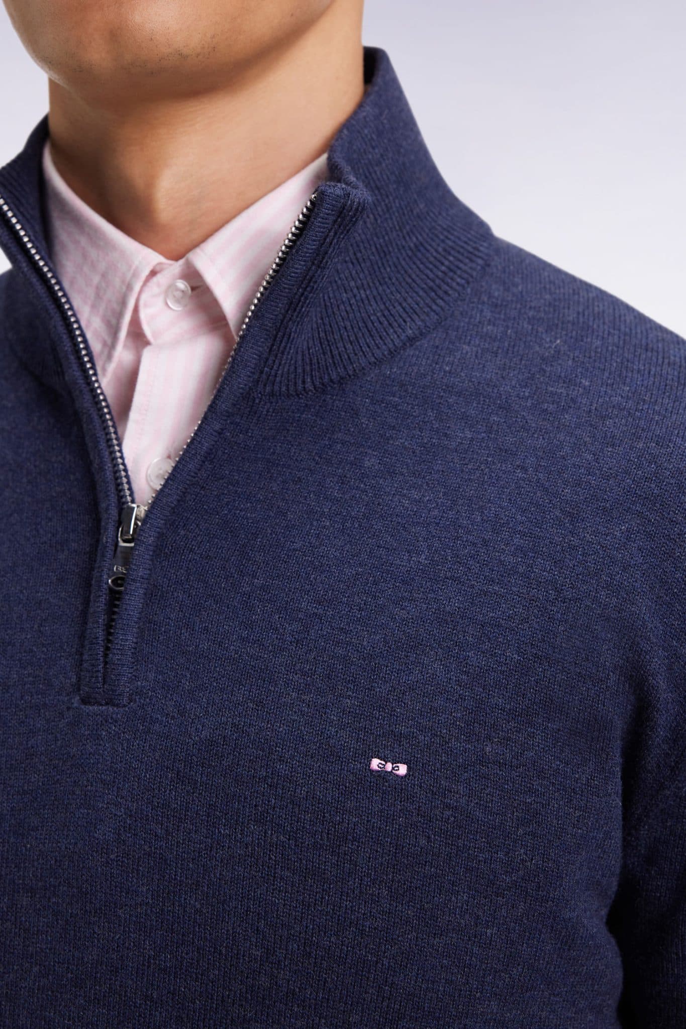 A person wearing a pale pink collared shirt under a navy blue, half-zip sweater with a small embroidered logo on the chest. Only the lower face, neck, and upper torso are visible.